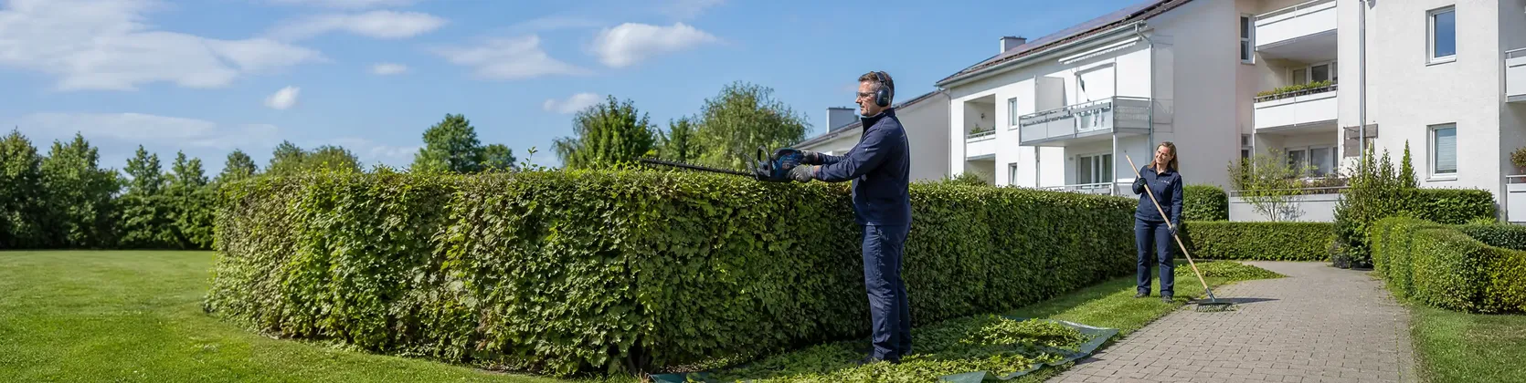 Mitarbeiter schneidet mit Schutzausrüstung eine Hecke an einer Wohnanlage, Kollegin recht Schnittgut zusammen