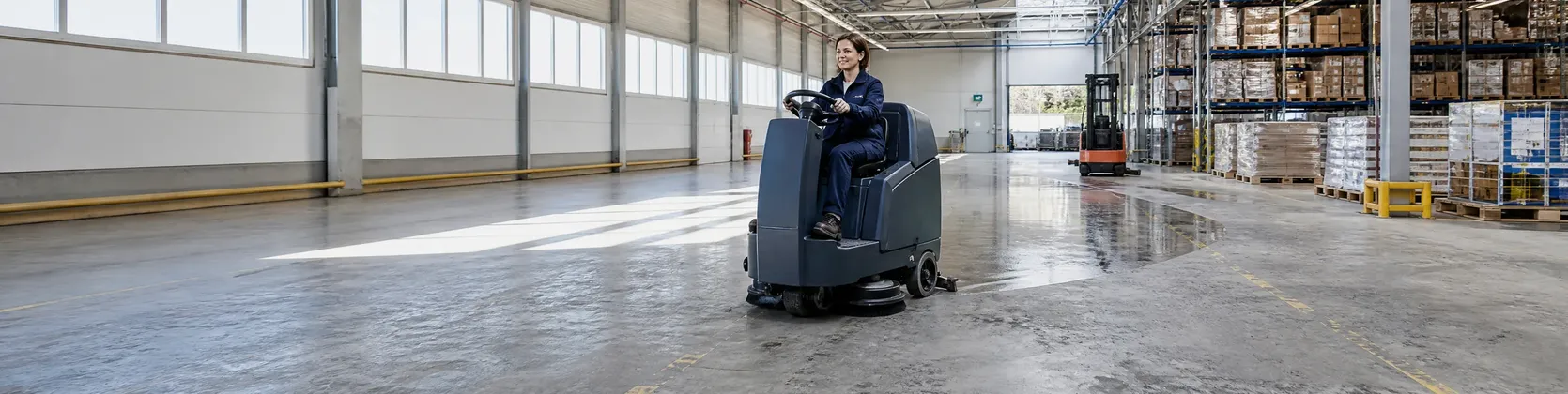 Mitarbeiterin reinigt mit einer Aufsitz-Scheuersaugmaschine den Boden einer Lagerhalle